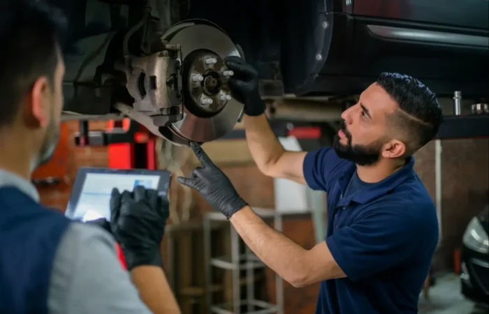 Brake replacement: A mechanic in black gloves pointing at a car's brake rotor while another technician records data on a tablet.