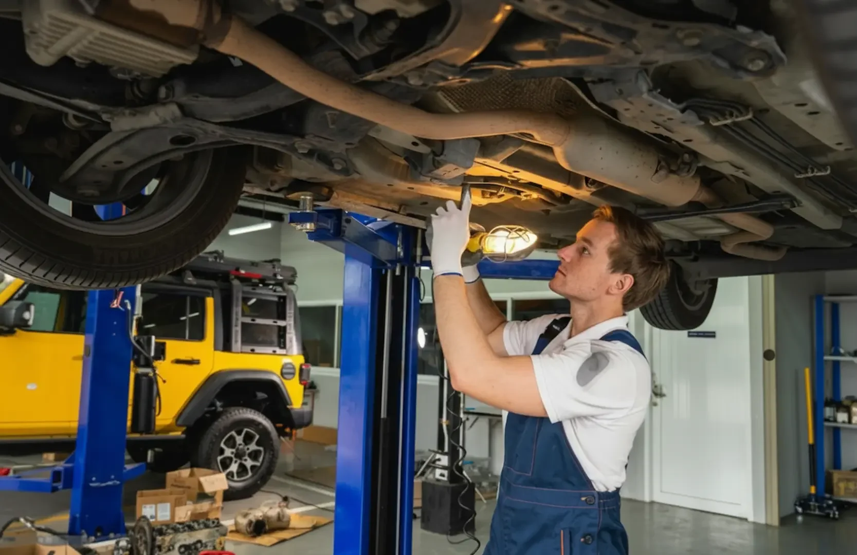 Brake Replacement: A technician standing under a vehicle on a lift, using a lamp to inspect the undercarriage and exhaust system.