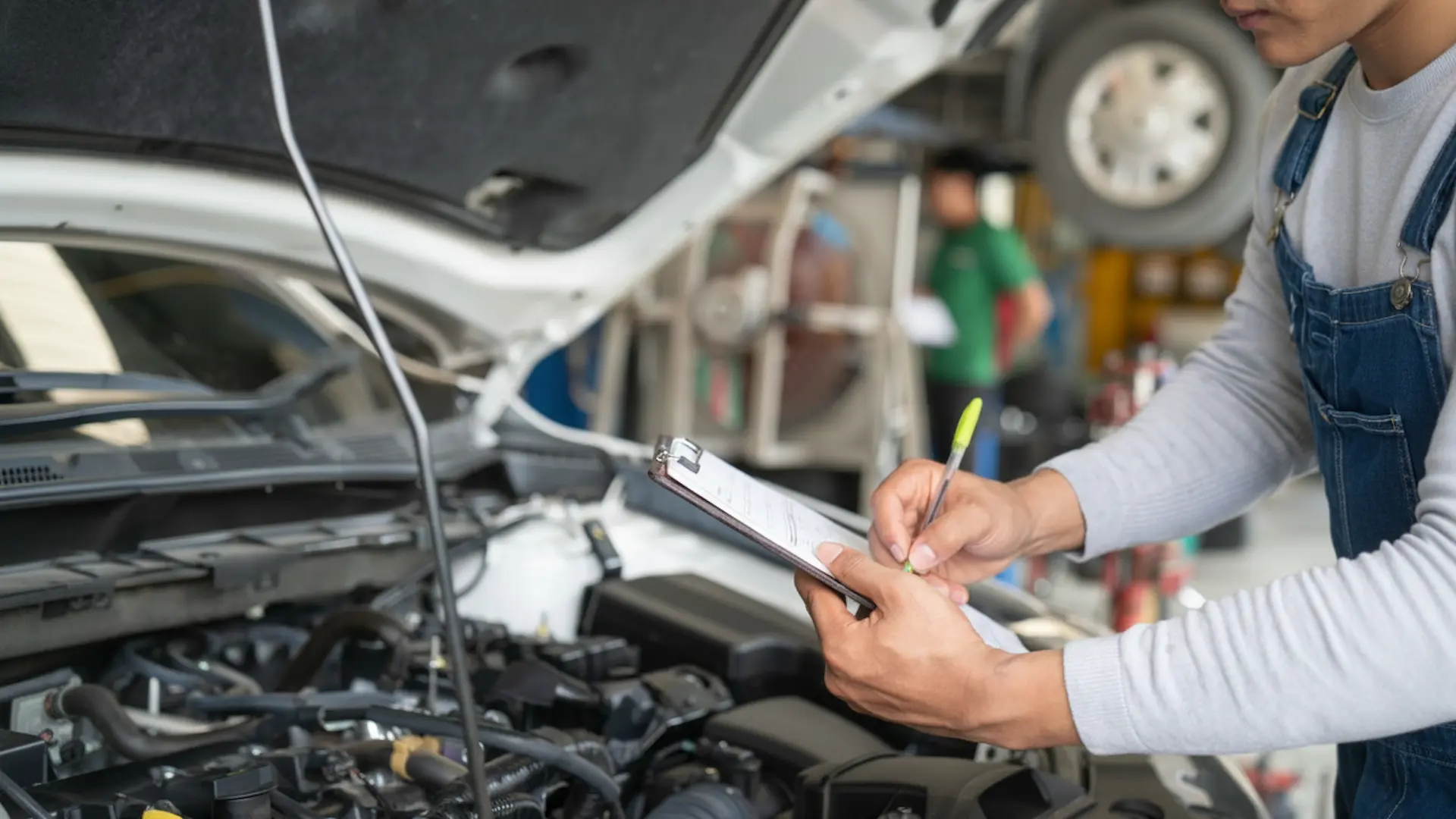 Vehicle Maintenance: A service adviser in denim overalls writing notes on a clipboard while standing by an open car hood.