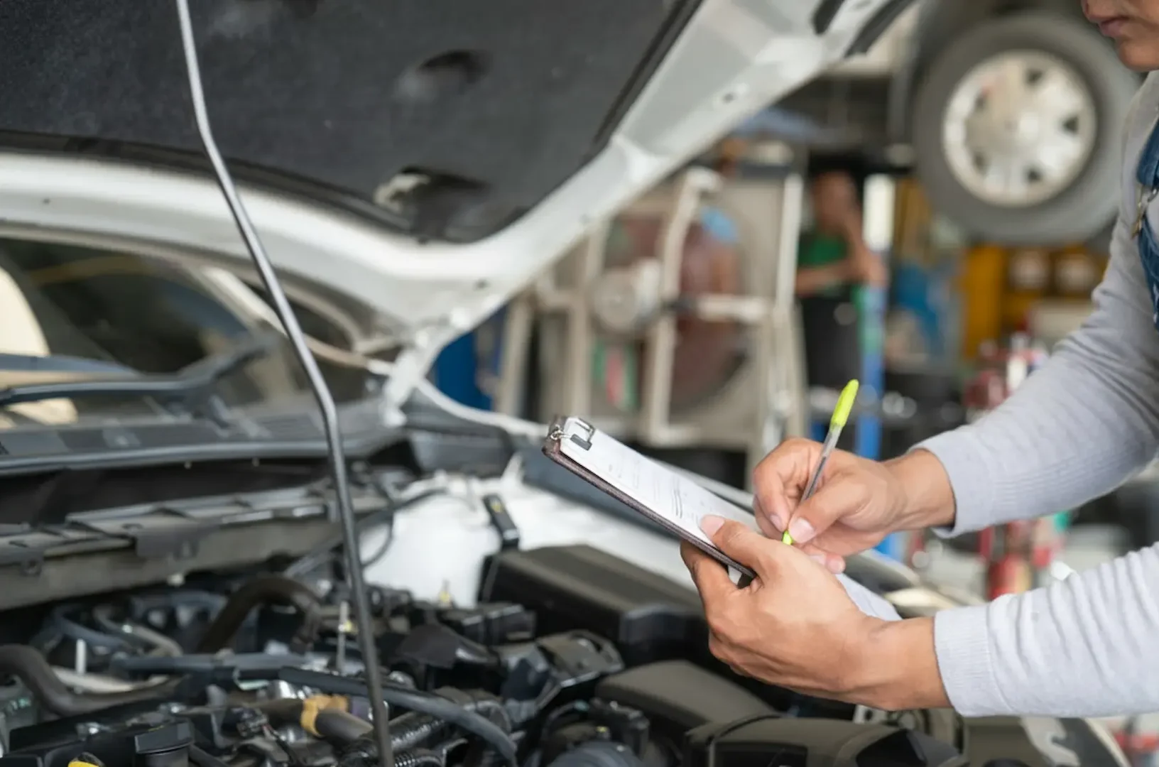 Close-up of expert Mechanics hands filling out a maintenance inspection checklist under the open hood of a car at Southport Auto Repair.