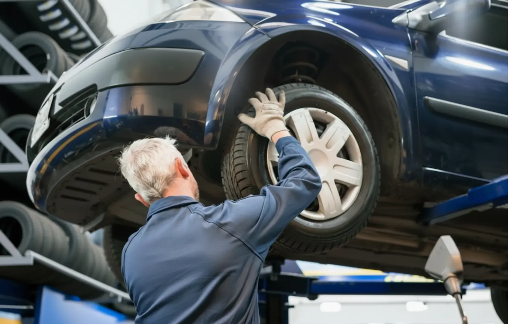 Affordable tire Deals: A technician in a blue workshop inspecting the tire and wheel assembly of a car raised on a hydraulic lift.