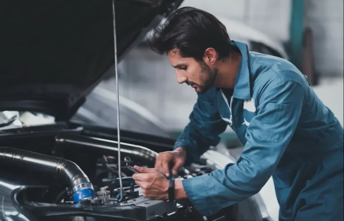 Vehicle Maintenance: A male auto technician in a blue uniform leaning over an open car hood to inspect engine components.