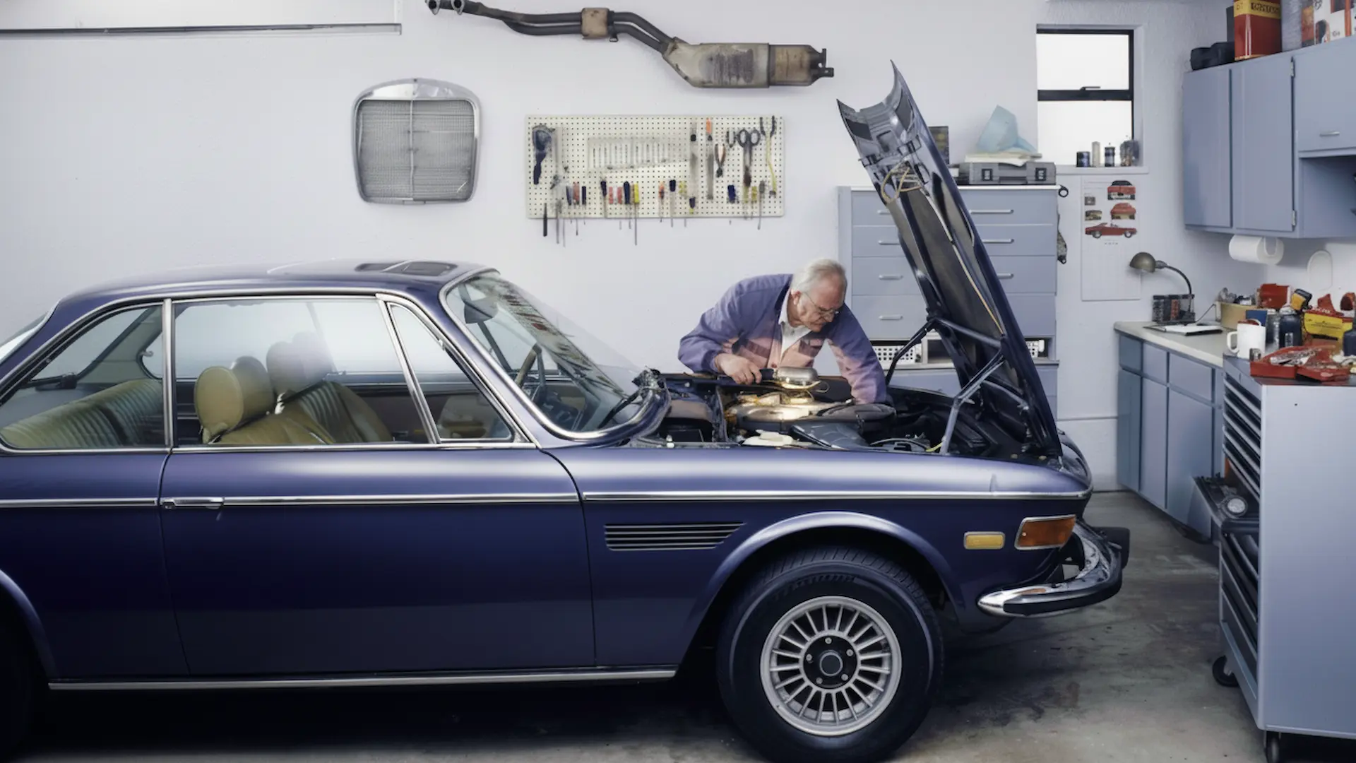 An elderly man performing engine maintenance on a vintage purple luxury coupe inside a well-equipped home workshop.