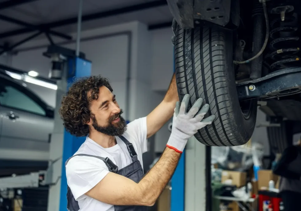 Auto repair mechanic wearing gloves and grey overalls inspecting a tire and suspension coil on a vehicle lift.