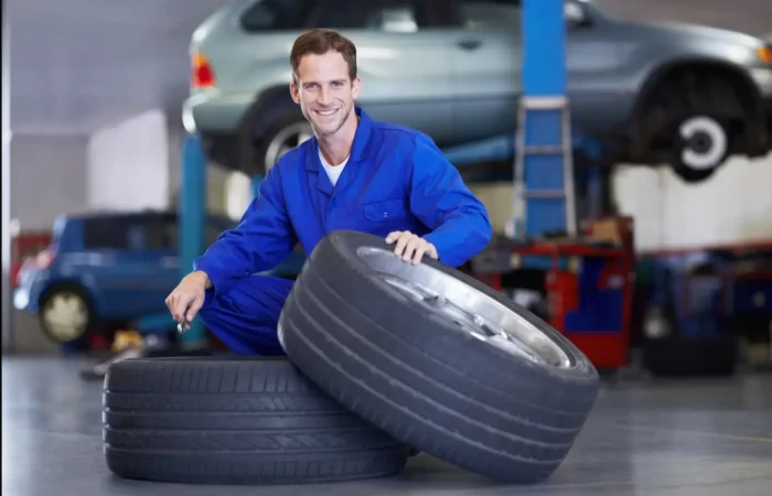 Best tire shops: A smiling mechanic in blue workwear kneeling next to a stack of new tires in a bright repair shop.