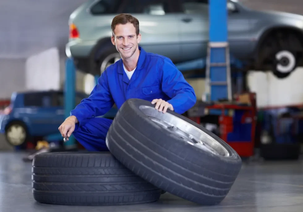 Best tire shops: A smiling mechanic in blue workwear kneeling next to a stack of new tires in a bright repair shop.