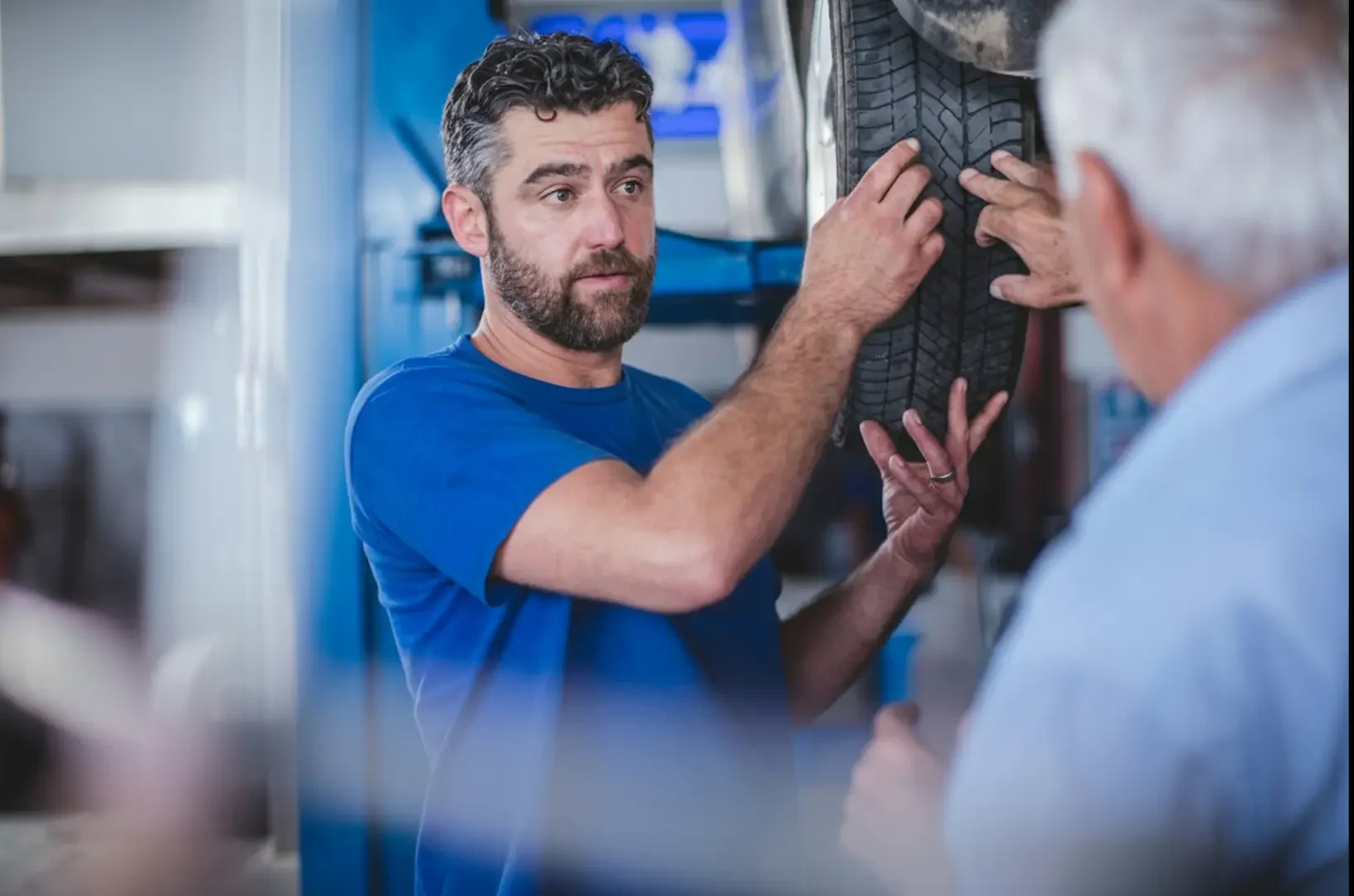 Auto repair mechanic in a blue shirt examining a tire on a lifted vehicle and discussing wear patterns.