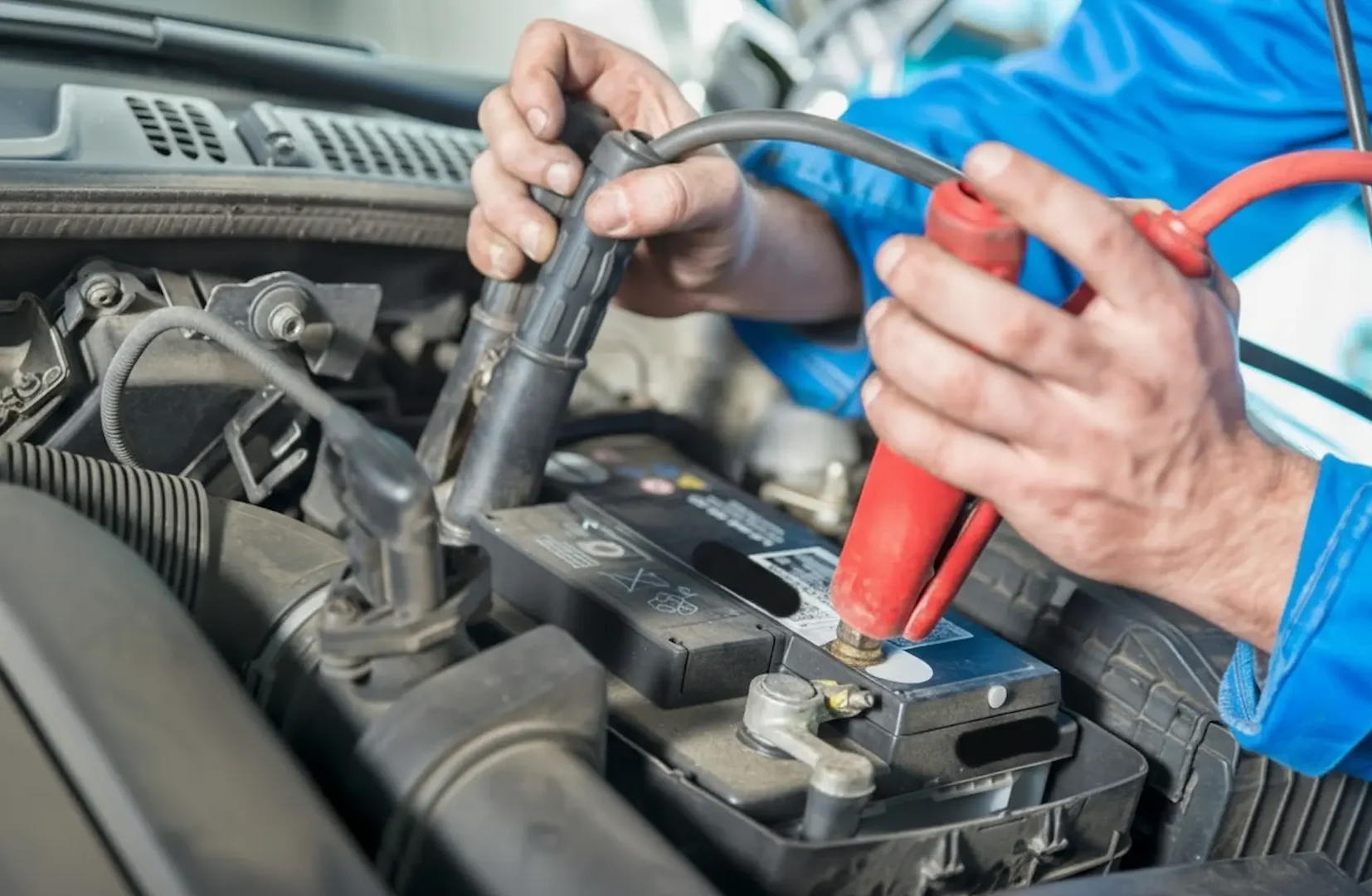 A technician in a blue uniform securely connecting red and black terminal clamps to a new car battery.