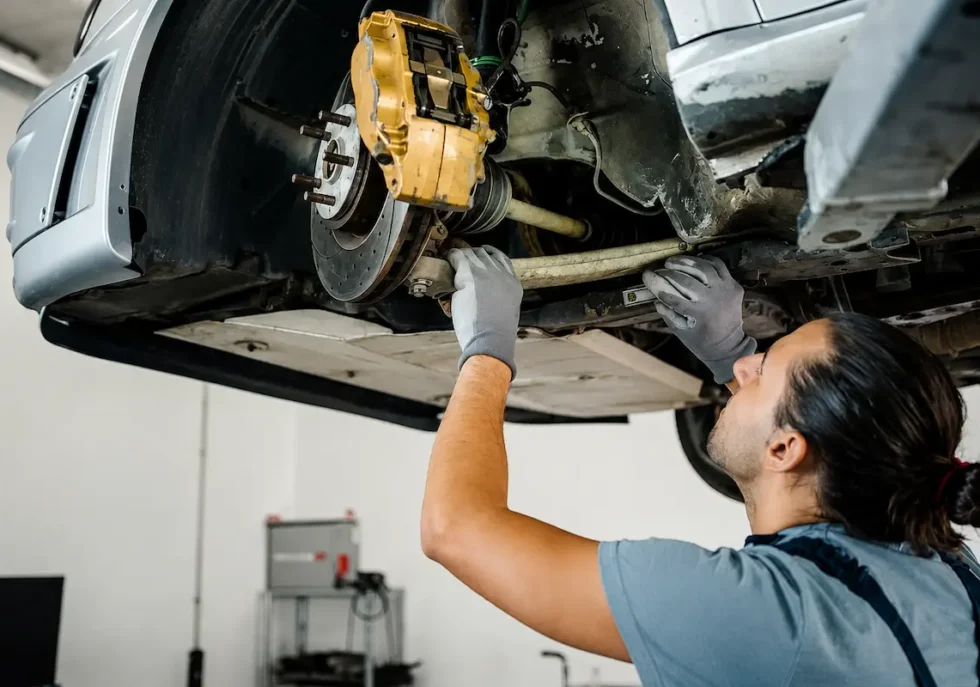 Mechanic performing suspension repair underneath a lifted car, inspecting brake and suspension components in an auto repair workshop.
